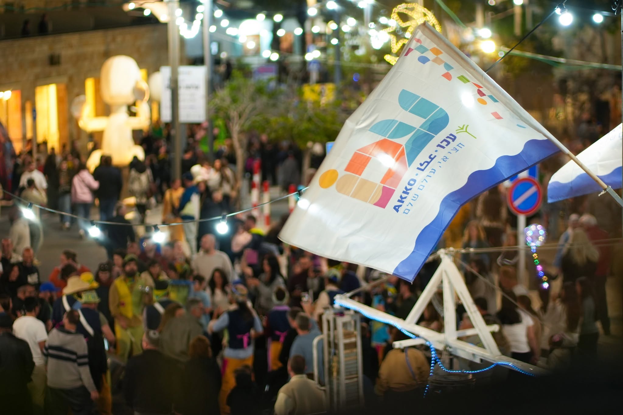 A night-time crowd in Acre beneath a festival flag raised above the stage.
