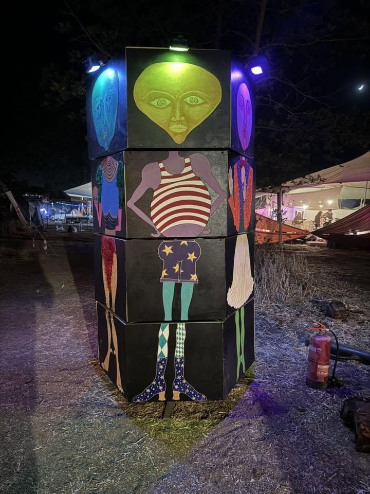 A green figure installation with a broad head glows between tents.