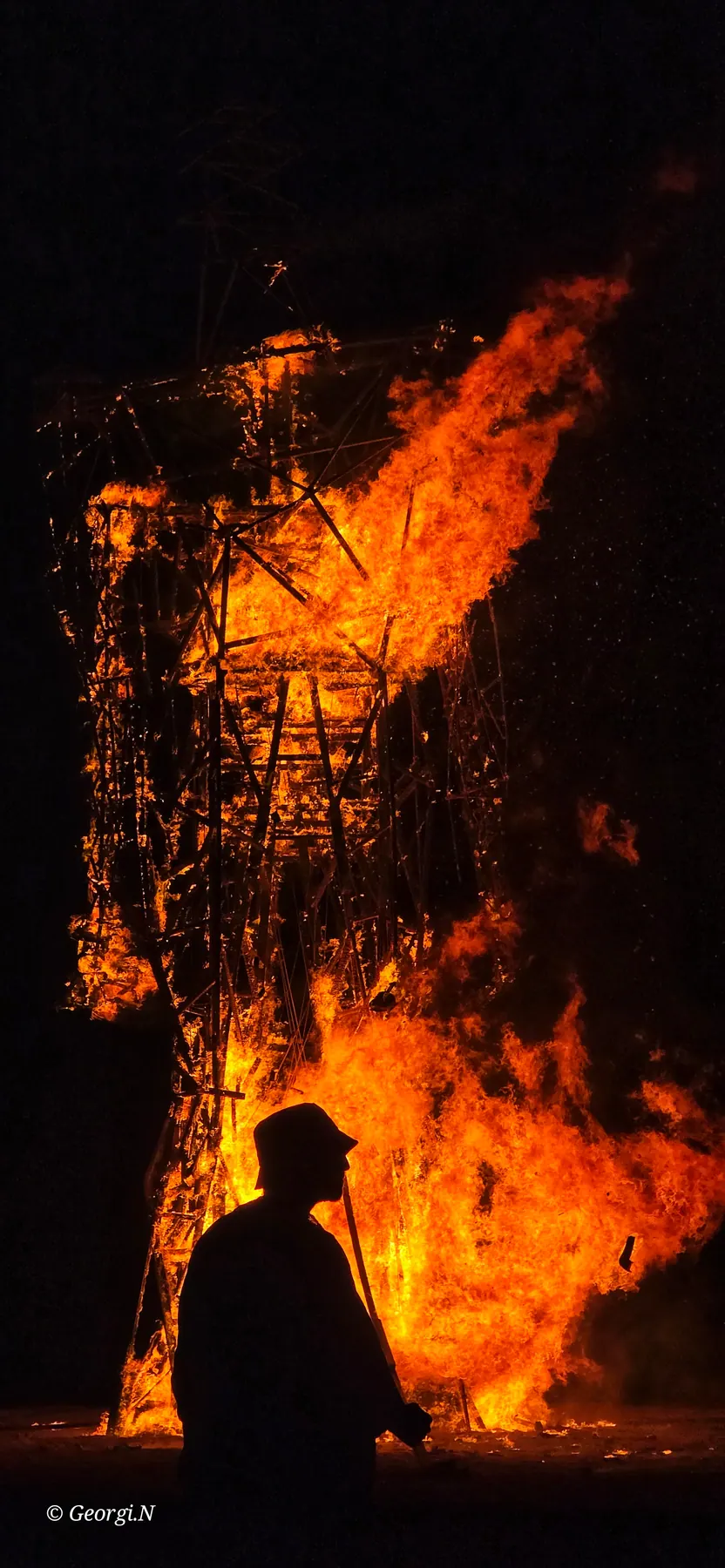 A person's silhouette stands before a burning wooden installation.