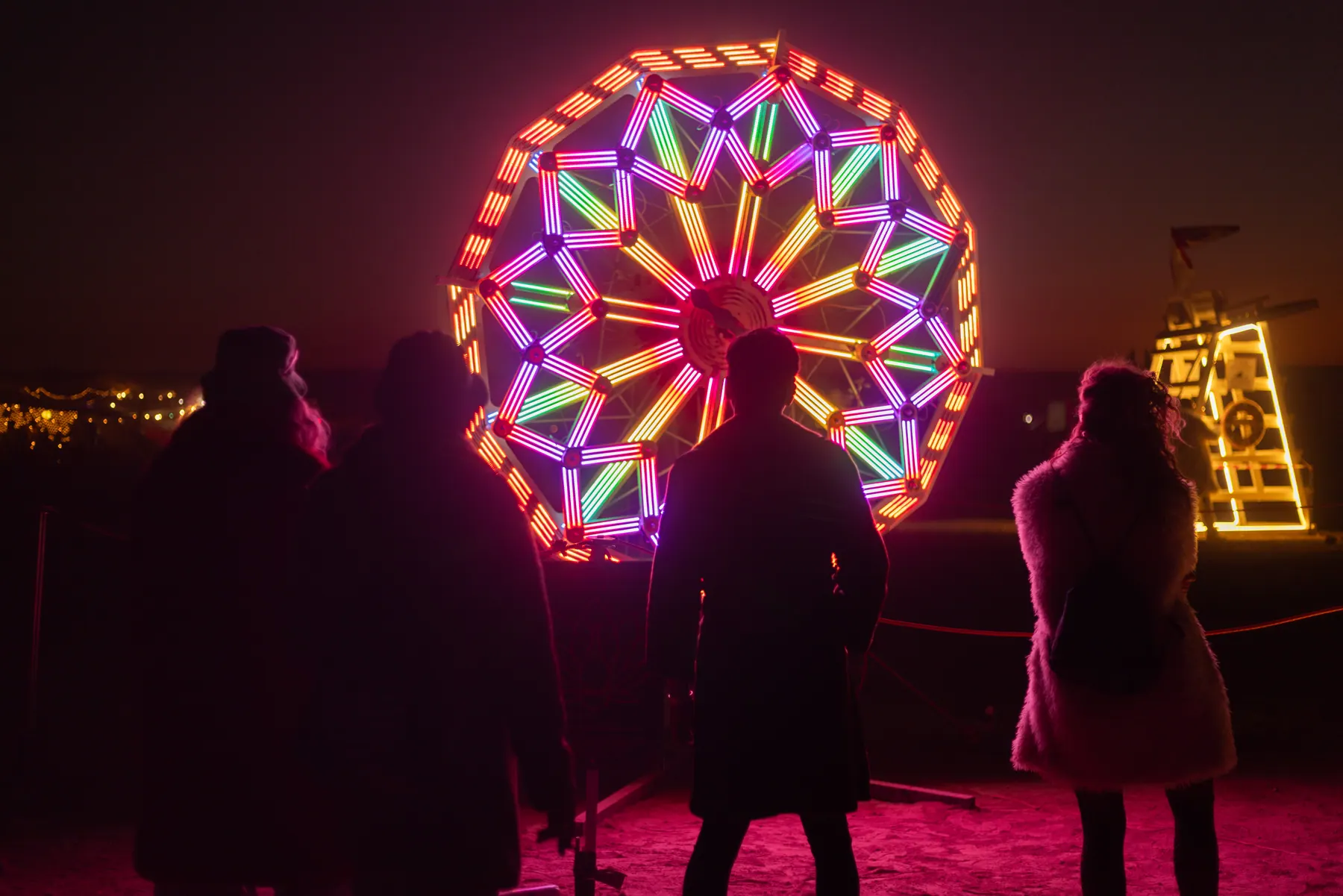 Three figures watch a colourful illuminated wheel at night.