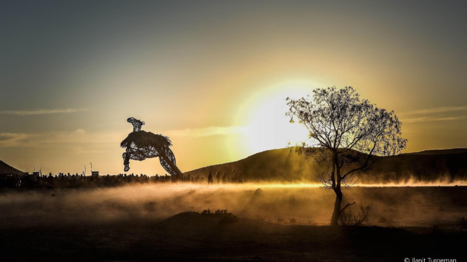 A silhouetted figure sculpture, a tree and drifting dust against the setting sun.