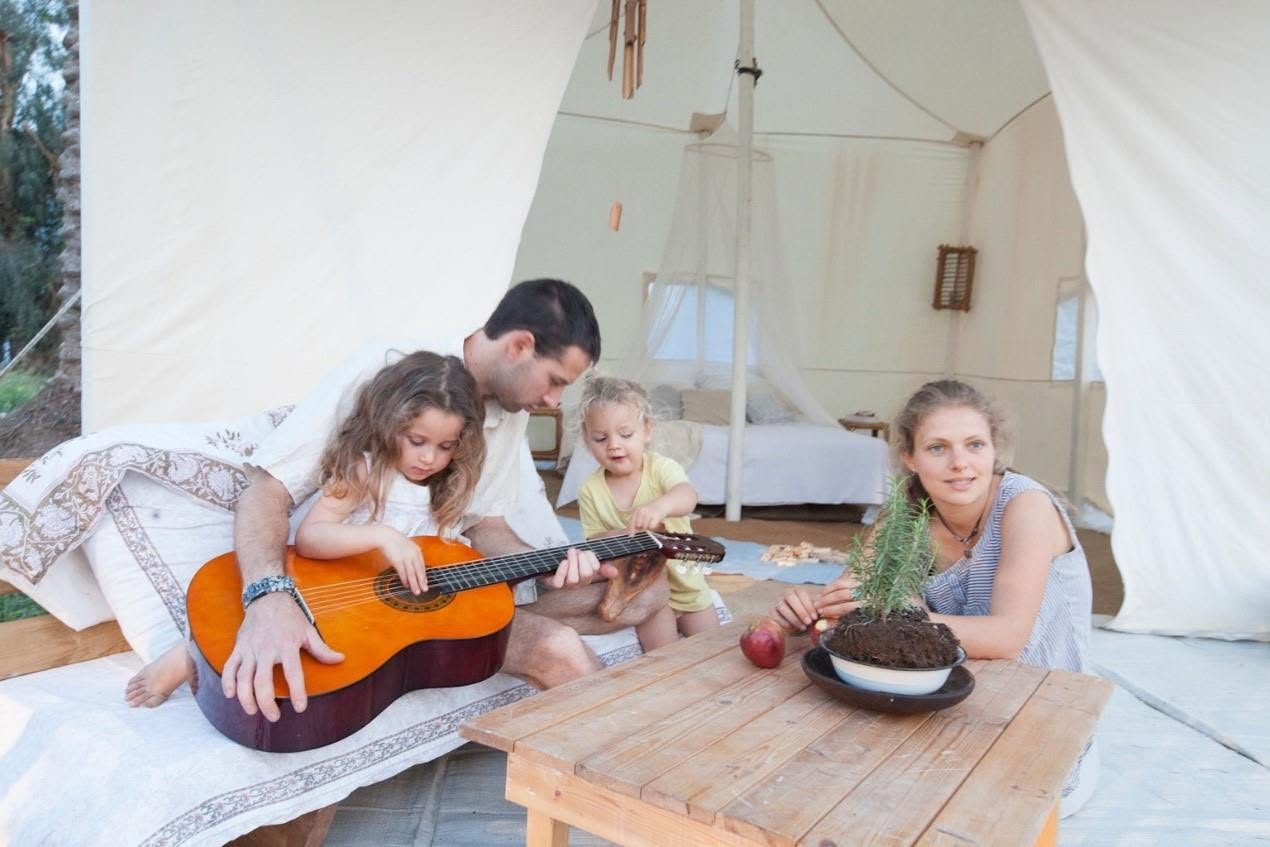 A family plays guitar around a small table inside a tent.